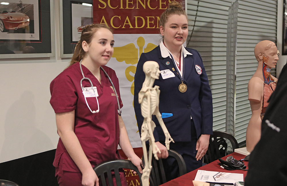 two students stand over a table with models of a human skeleton and other medical models