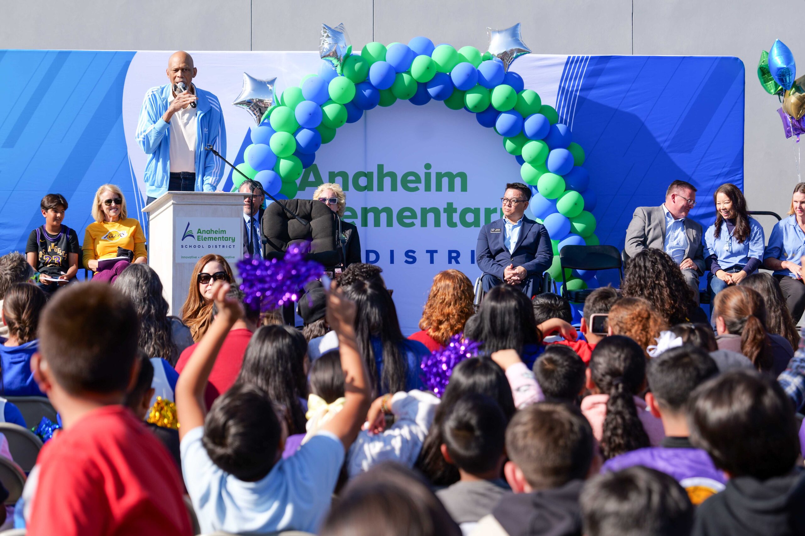 NBA legend Kareem Abdul-Jabbar speaks to a crowd of students and educators during a daytime outdoor assembly at Thomas Edison Elementary School. He stands at a podium in front of a balloon arch and Anaheim Elementary School District signage, flanked by seated guests including County Superintendent Dr. Stefan Bean.