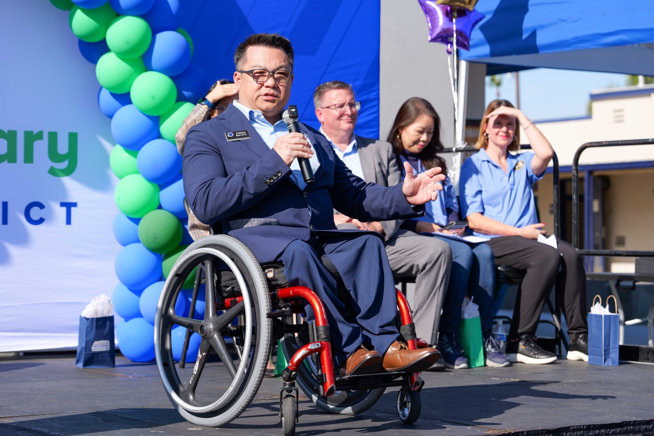 Dr. Stefan Bean, Orange County superintendent of schools, speaking on an outdoor stage decorated with green and blue balloons. Four panelists sit behind him.