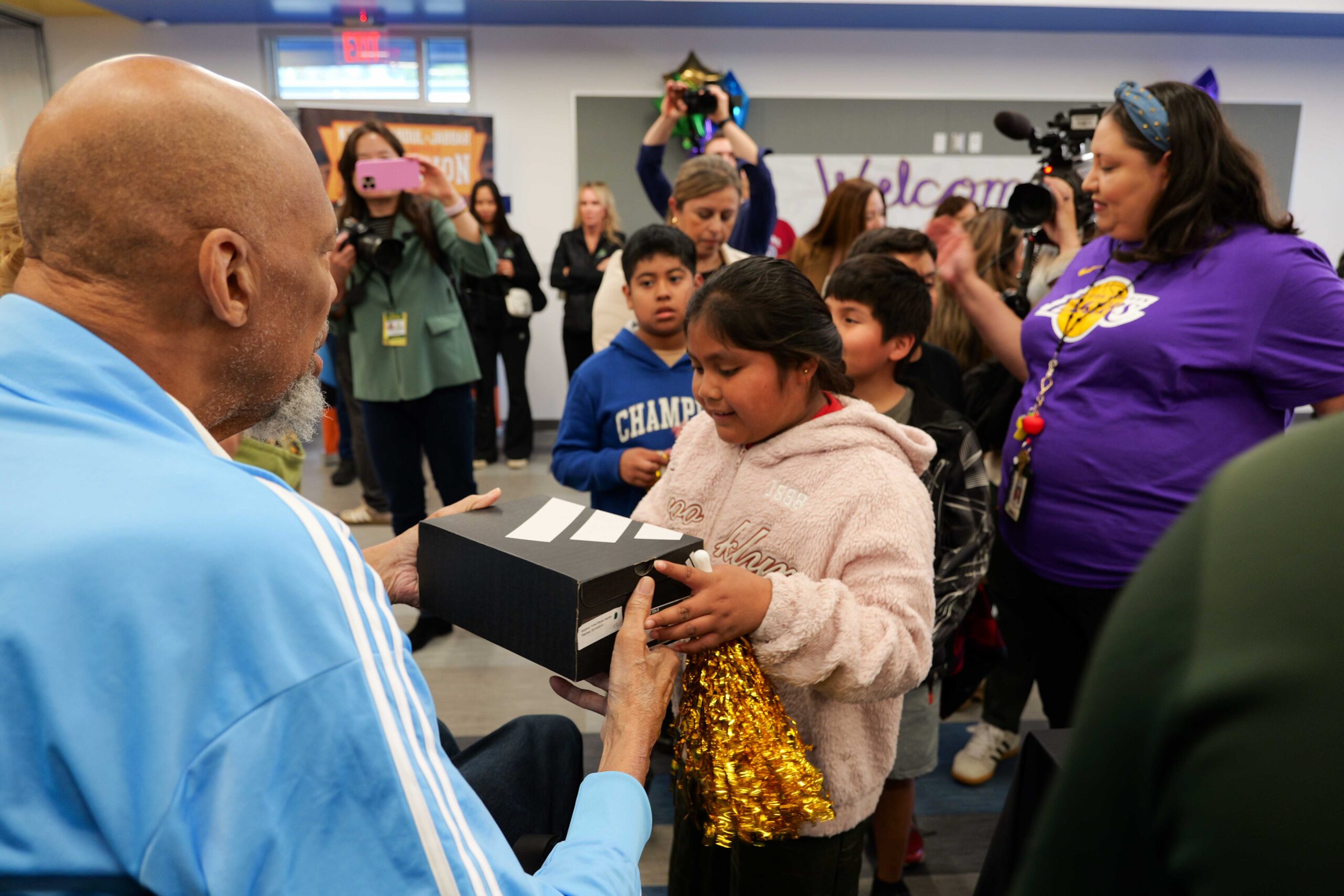 Kareem Abdul-Jabbar hands a student a shoebox as she smiles and accepts it, surrounded by classmates and educators in a classroom setting.