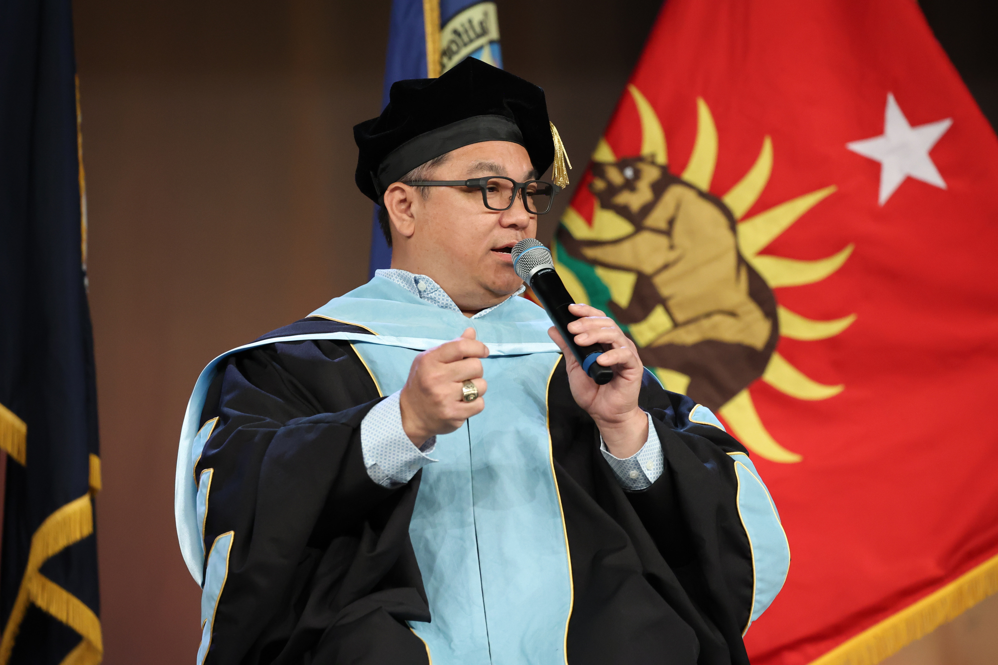 Dr. Stefan Bean speaks at the podium in academic regalia