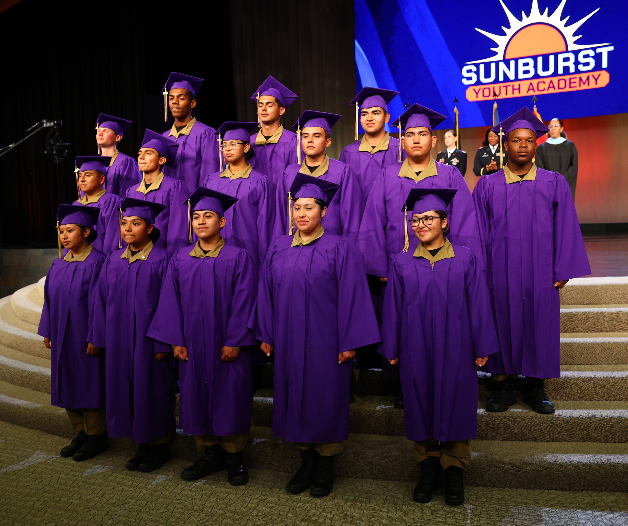 Sunburst Youth Academy graduates pose on stage in purple caps and gowns