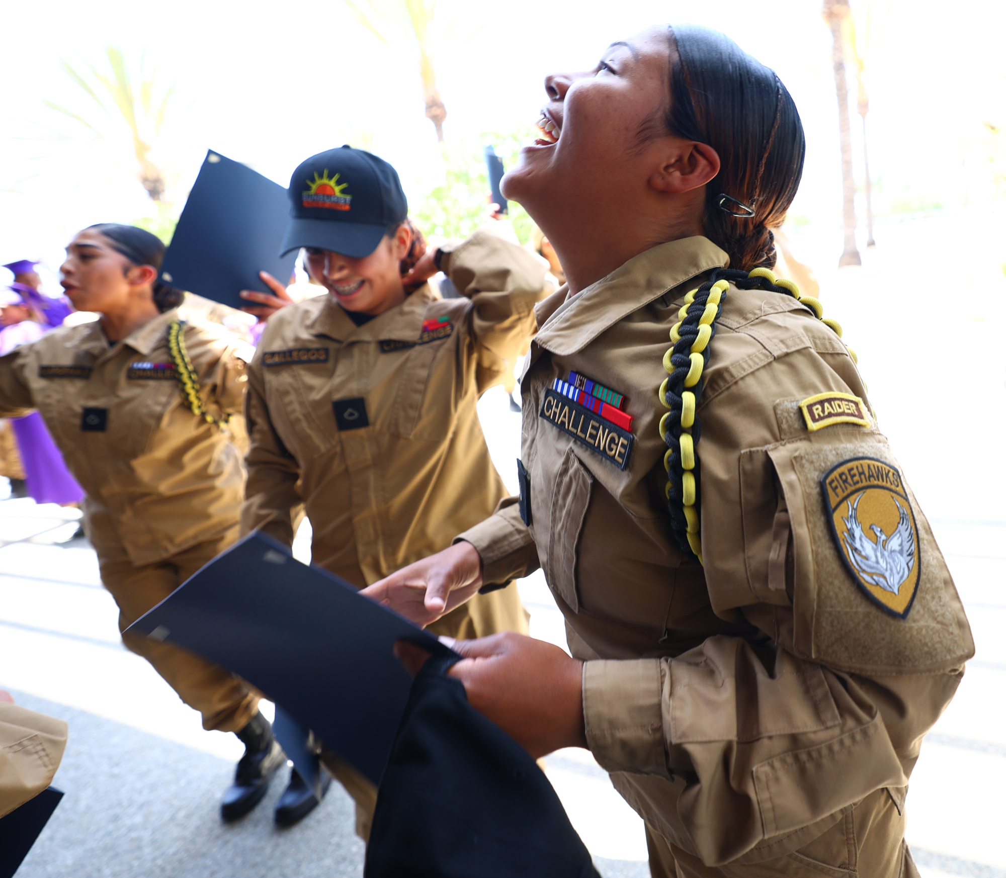Cadets from Sunburst Youth Academy in uniform smile and laugh while holding diplomas and celebrating graduation outdoors.