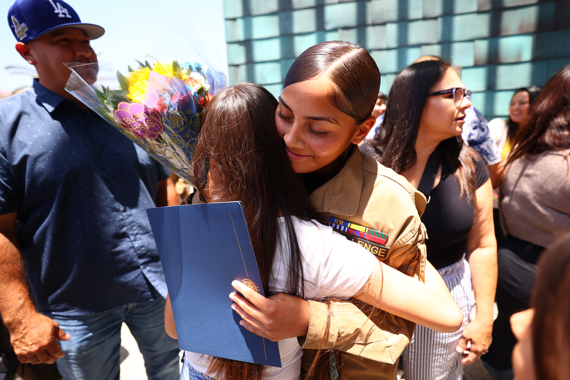 A female cadet in uniform hugs a young girl holding a bouquet and diploma, with family members smiling nearby.