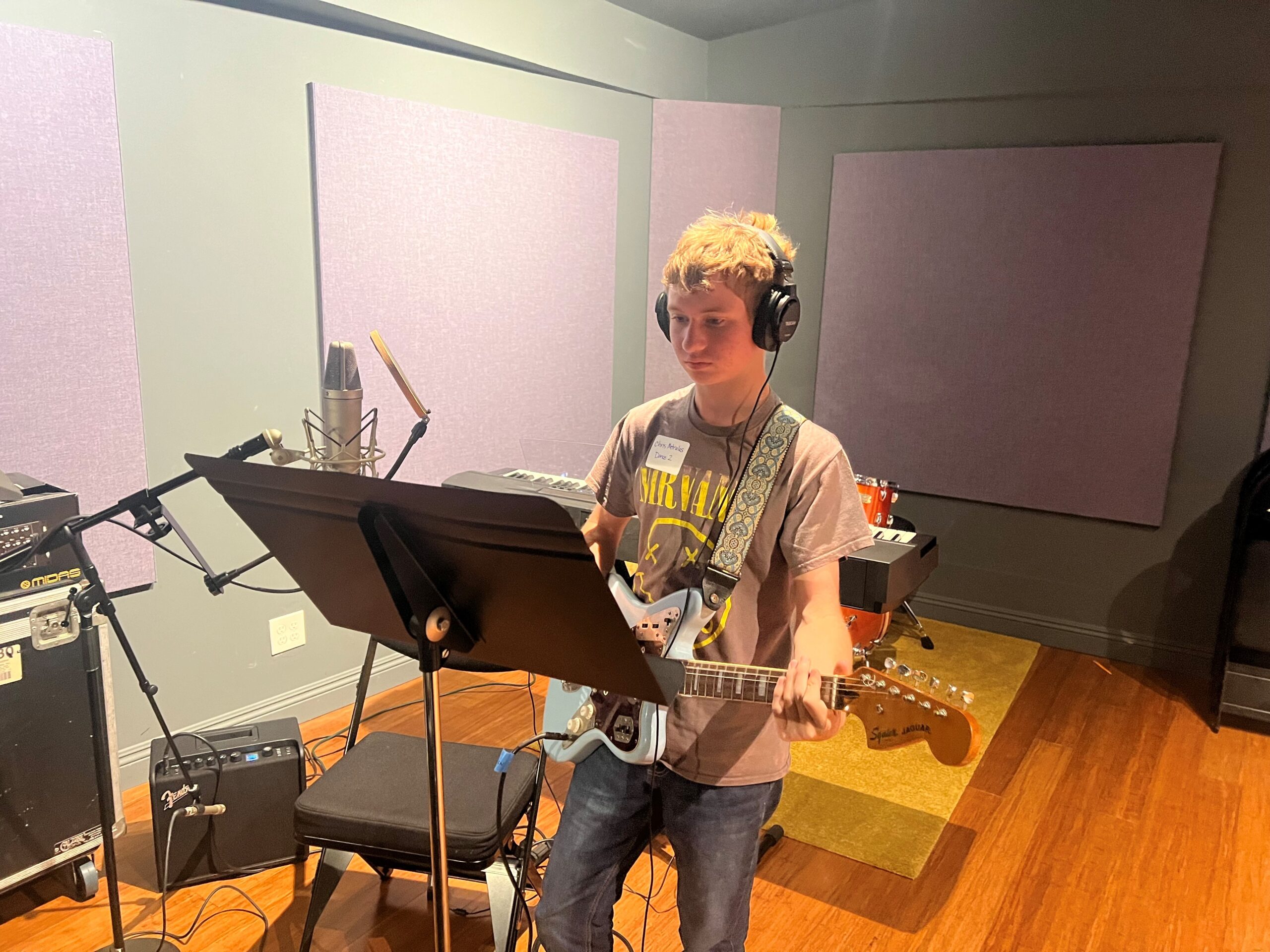 A high school student wearing headphones plays an electric guitar and stands at a microphone in a recording studio during the Fender Play songwriting workshop.