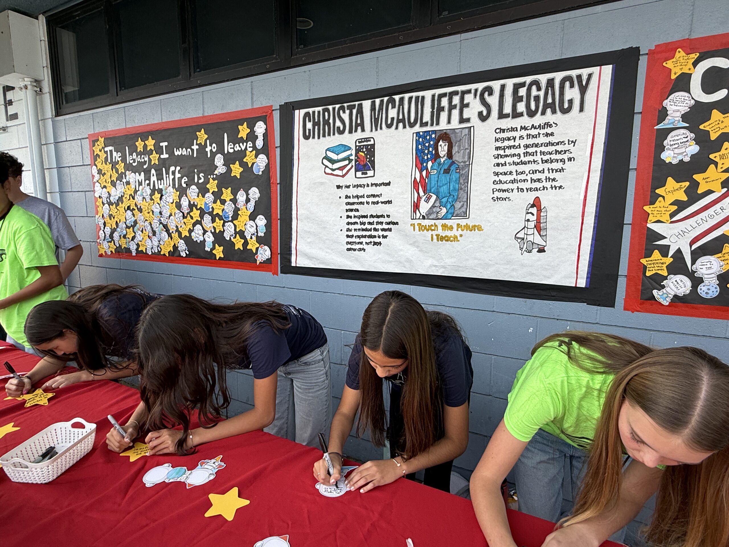 Students at McAuliffe Middle School write reflections on paper stars during a campus activity honoring Christa McAuliffe’s legacy, with student-created posters displayed behind them.