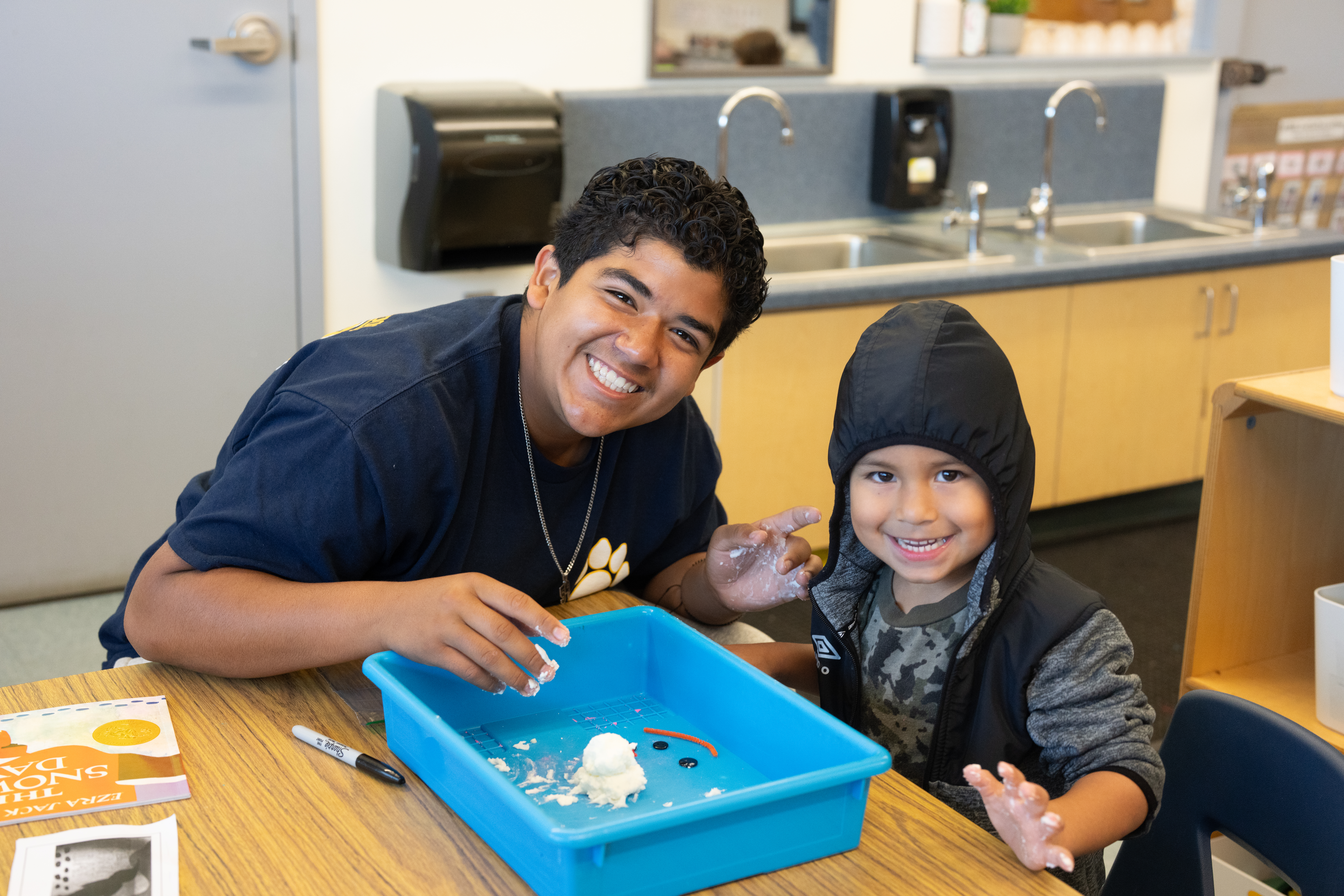 An older student smiles with a transitional kindergarten student while working on an activity together after reading.