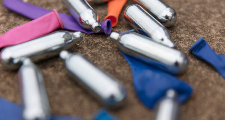 Scattered nitrous oxide canisters and colorful balloons on pavement, often used for recreational inhalation.
