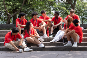 High school students in red T-shirts sitting on stone steps, engaged in conversation during a leadership program event, with trees in the background