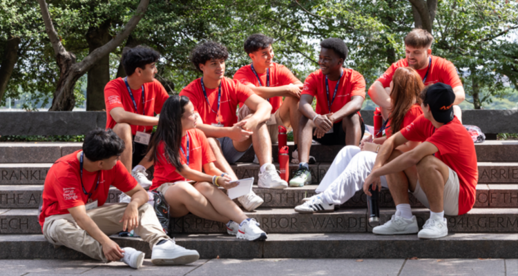 High school students in red T-shirts sitting on stone steps, engaged in conversation during a leadership program event, with trees in the background