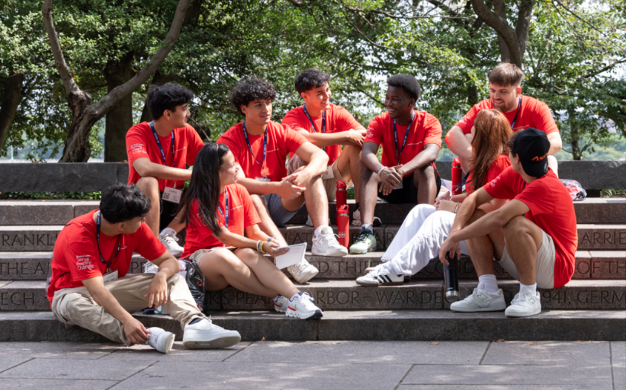 High school students in red T-shirts sitting on stone steps, engaged in conversation during a leadership program event, with trees in the background