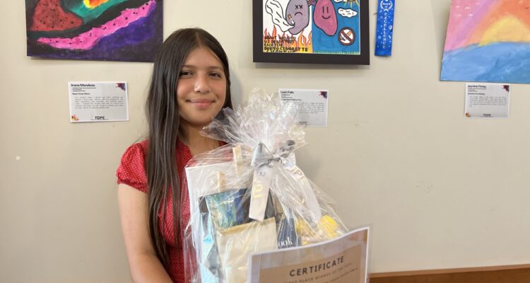 Leah Marie Felix holding a gift basket and certificate in front of her winning artwork — a colorful drawing of lungs promoting a tobacco-free lifestyle — at the 2025 TUPE Art Showcase.
