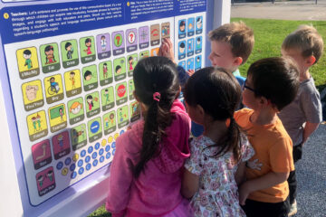 A group of elementary school students stands at a communication board outdoors, interacting with symbols and images together.