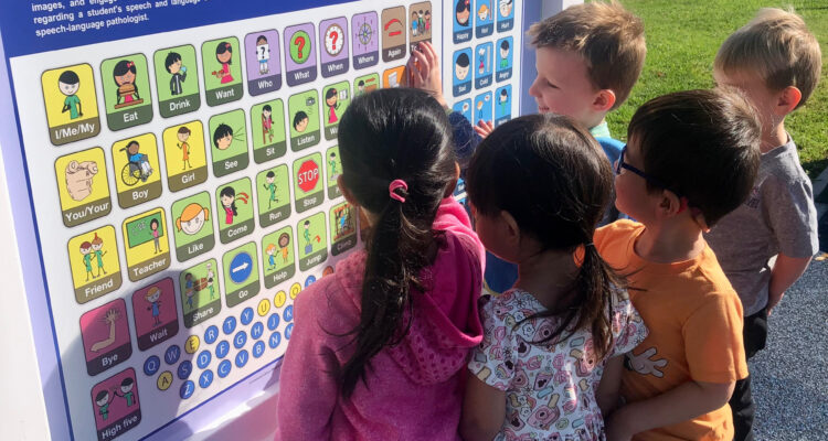 A group of elementary school students stands at a communication board outdoors, interacting with symbols and images together.