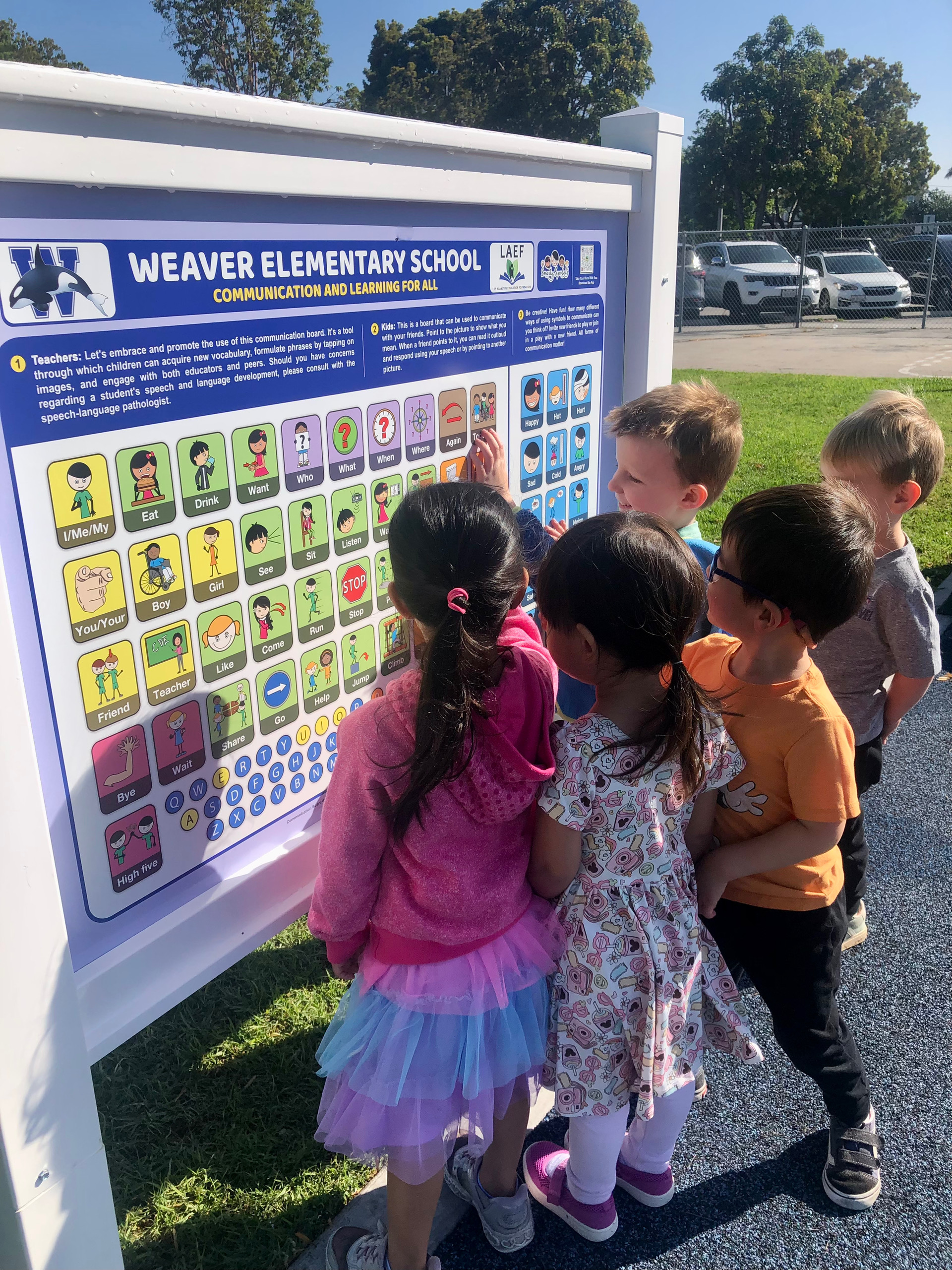 A group of elementary school students stands at a communication board outdoors, interacting with symbols and images together.