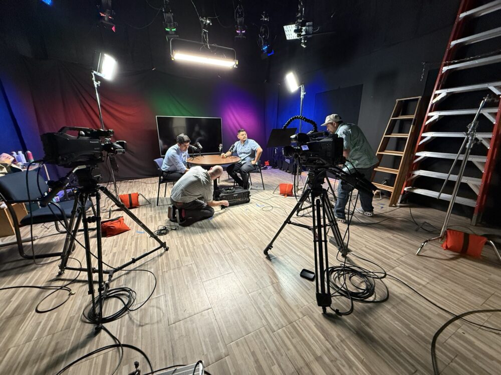 Studio crew adjusting cameras, lighting and sound equipment while two people sit at a table under stage lights.