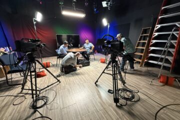 Studio crew adjusting cameras, lighting and sound equipment while two people sit at a table under stage lights.