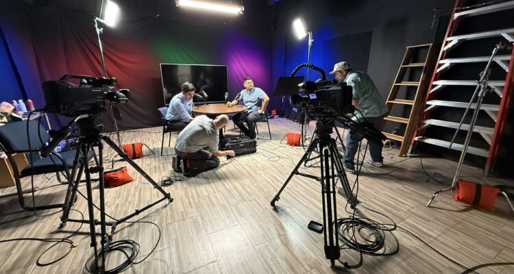 Studio crew adjusting cameras, lighting and sound equipment while two people sit at a table under stage lights.