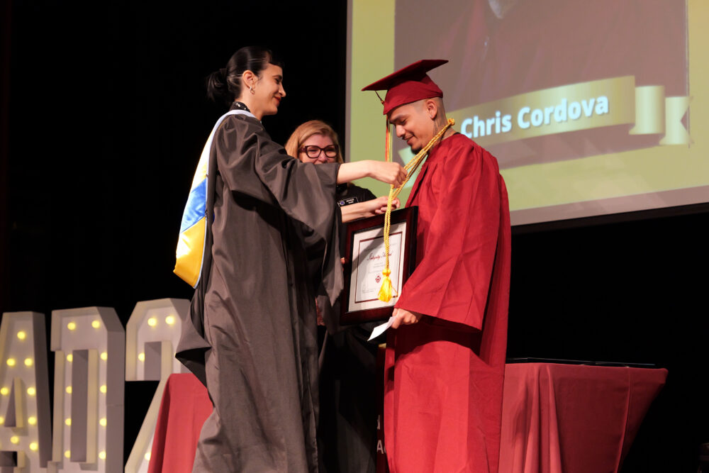 A graduate in a red cap and gown receives a medal and framed certificate on stage during CCPA’s June 2025 graduation ceremony