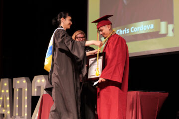 A graduate in a red cap and gown receives a medal and framed certificate on stage during CCPA’s June 2025 graduation ceremony