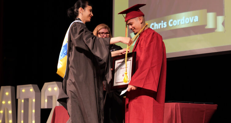 A graduate in a red cap and gown receives a medal and framed certificate on stage during CCPA’s June 2025 graduation ceremony