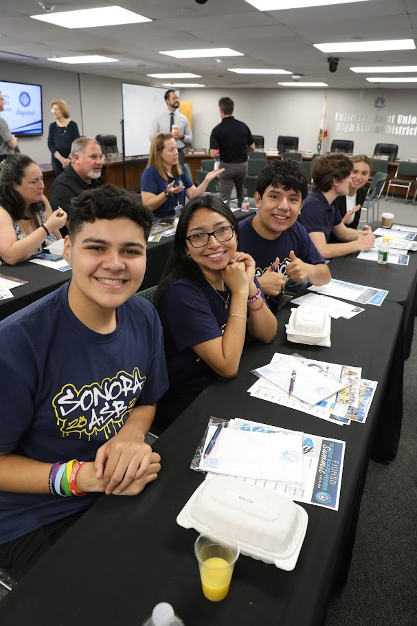 Three students wearing Sonora High School ASB shirts are seated at a table with papers, pens and food containers 