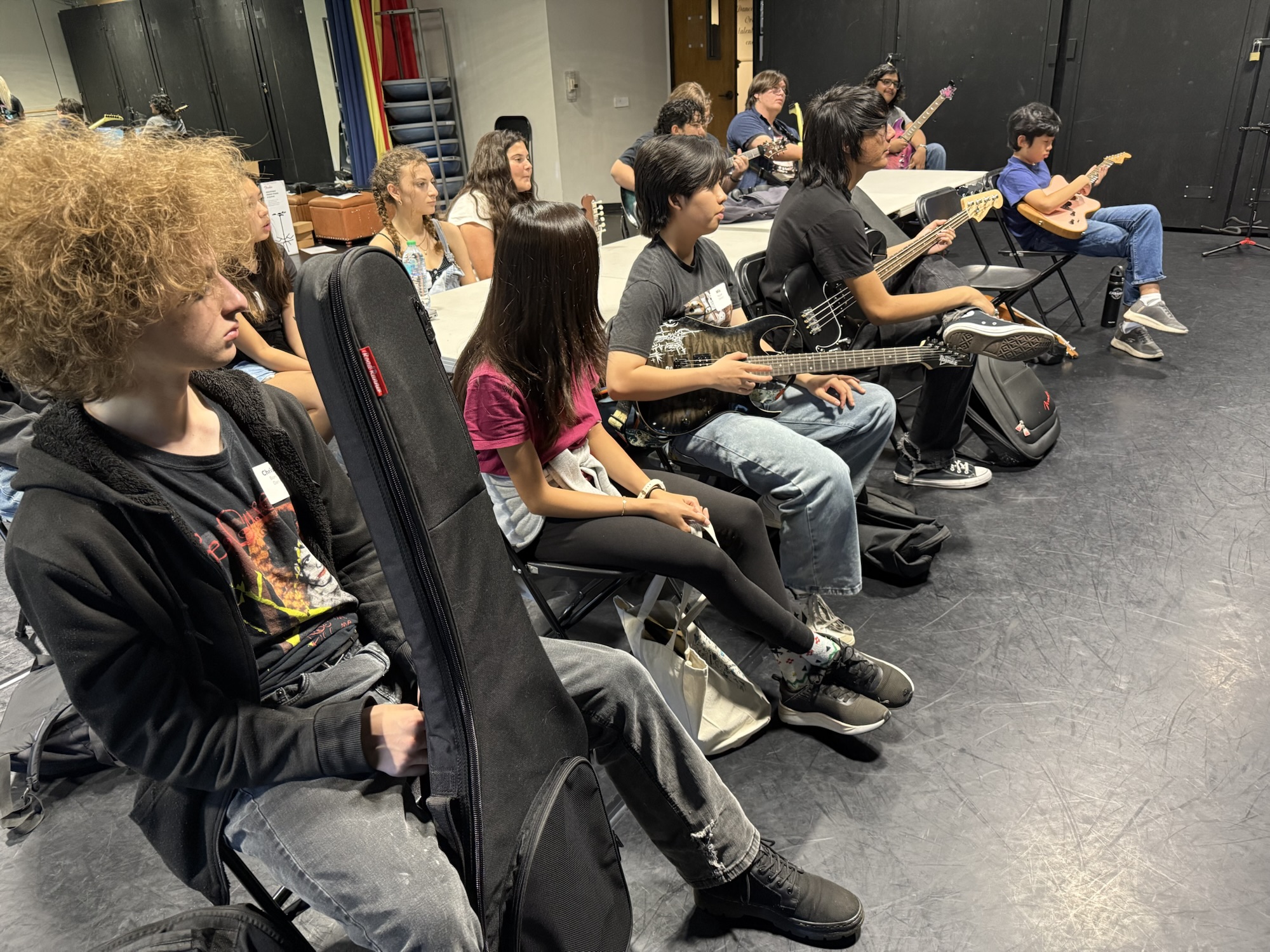 A group of high school students sit in chairs holding guitars and basses during a workshop session at OC Music & Dance.