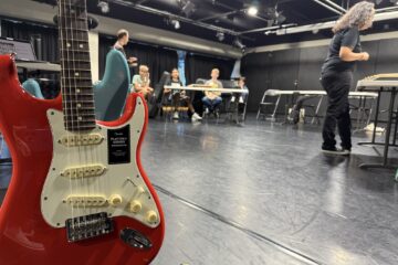 A red Fender Stratocaster guitar is pictured in the foreground as high school students sit at tables in a studio during the Fender Play songwriting workshop.
