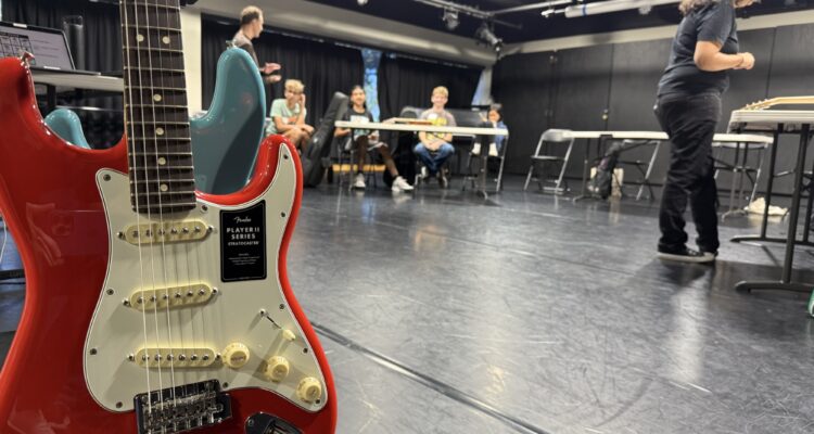 A red Fender Stratocaster guitar is pictured in the foreground as high school students sit at tables in a studio during the Fender Play songwriting workshop.