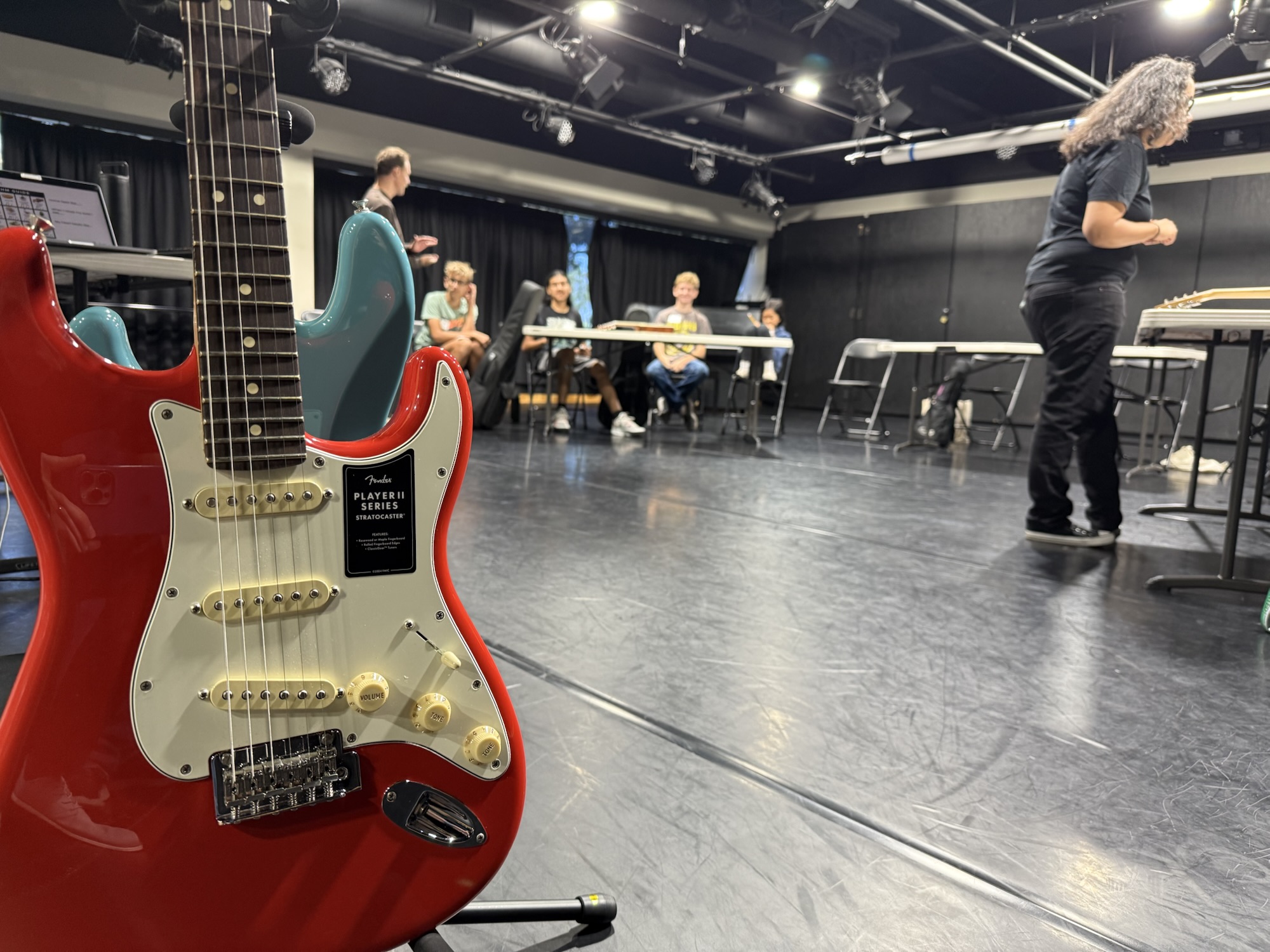 A red Fender Stratocaster guitar is pictured in the foreground as high school students sit at tables in a studio during the Fender Play songwriting workshop.