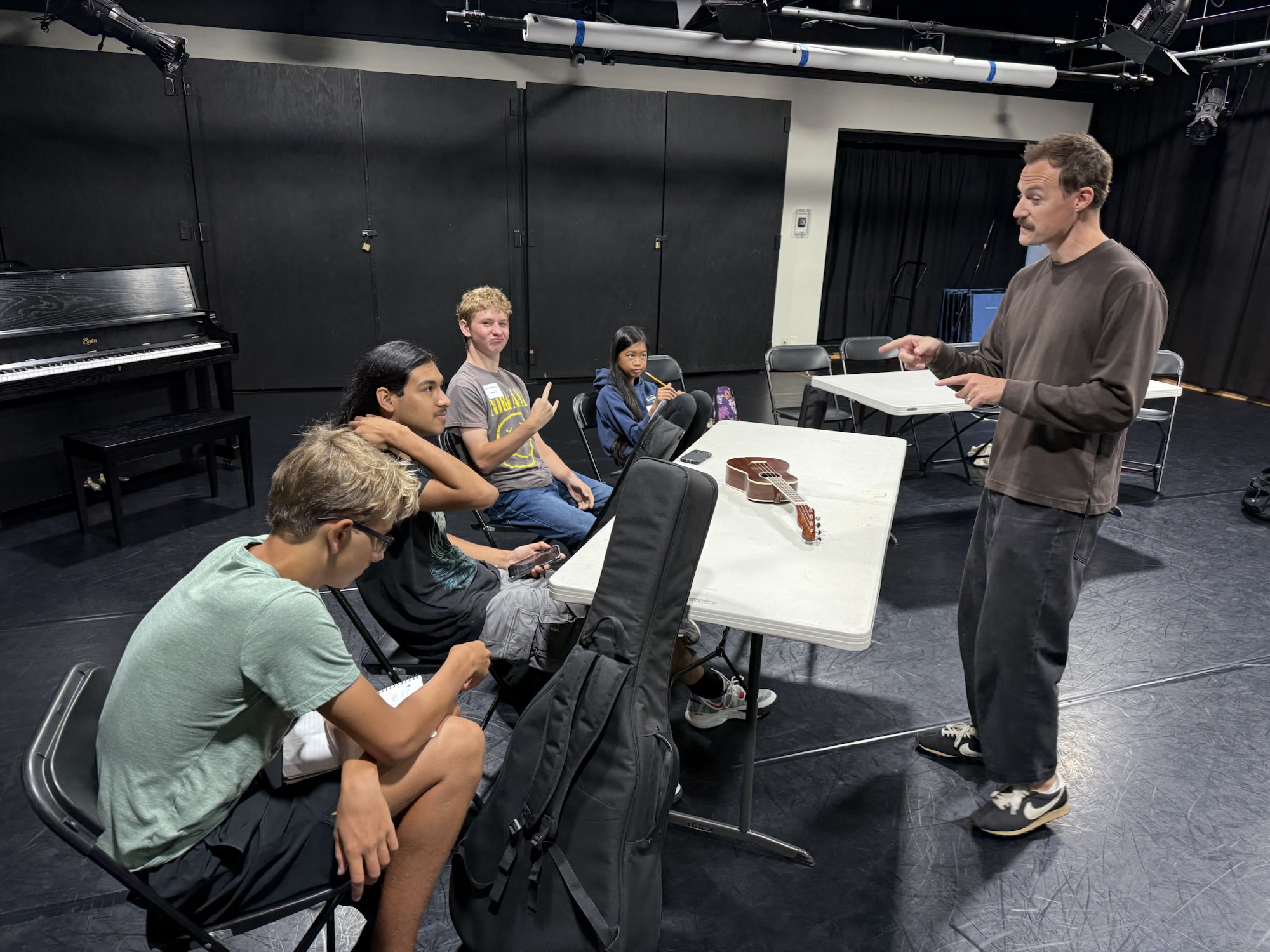 A music instructor speaks to a small group of high school students seated at tables in a black box studio at OC Music & Dance. A guitar rests on the table as the students listen and take notes.
