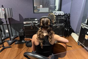 A high school student wearing headphones and holding an acoustic guitar sits at a microphone in a recording studio during the Fender Play songwriting workshop.
