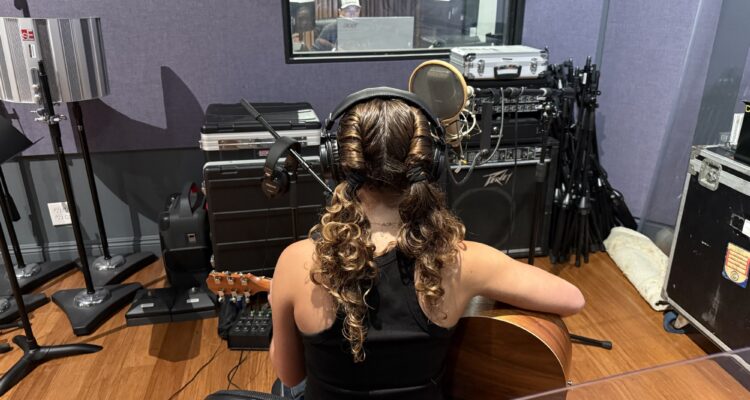 A high school student wearing headphones and holding an acoustic guitar sits at a microphone in a recording studio during the Fender Play songwriting workshop.