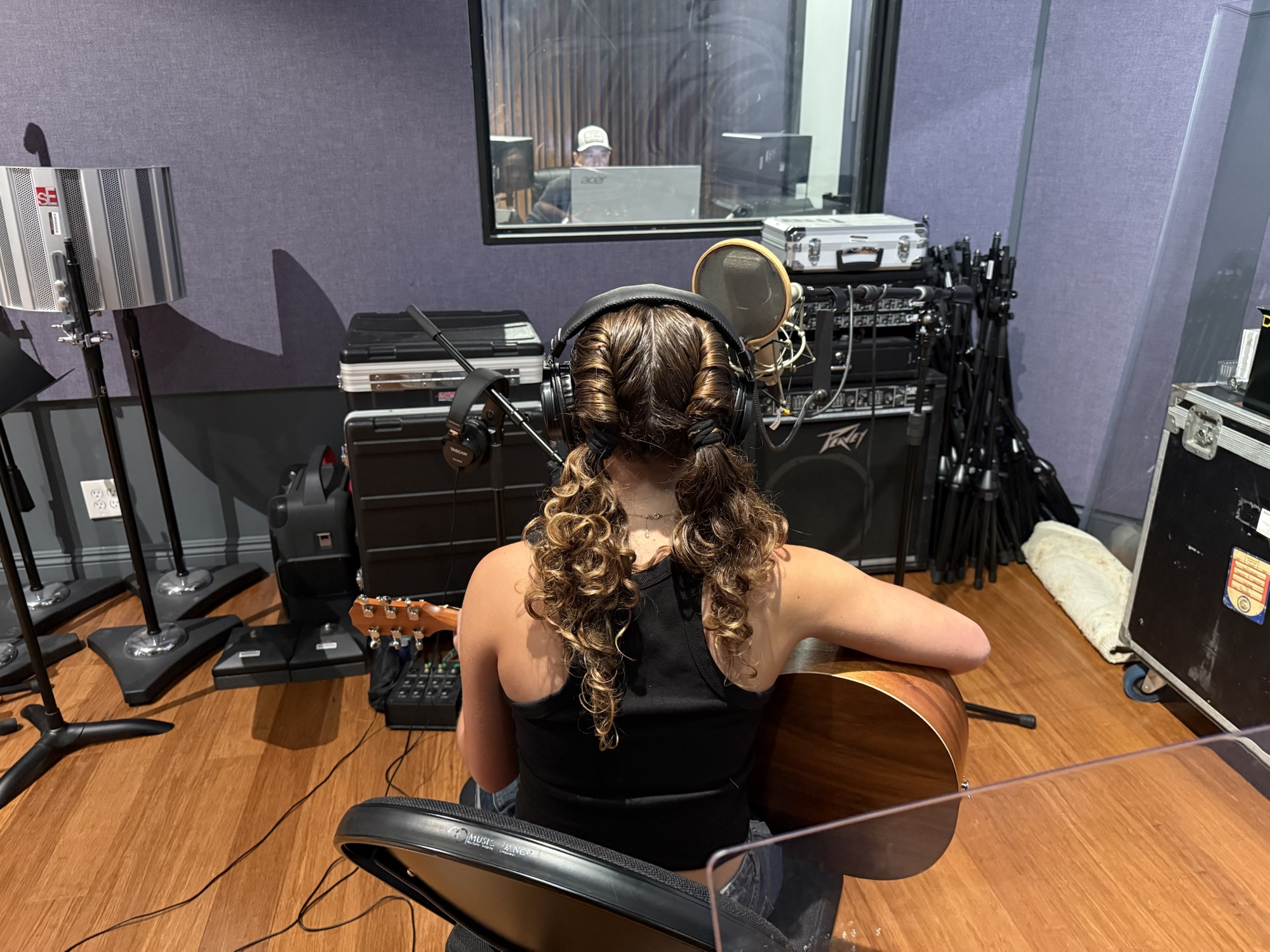 A high school student wearing headphones and holding an acoustic guitar sits at a microphone in a recording studio during the Fender Play songwriting workshop.