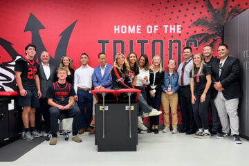 Group of San Clemente High School student-athletes and representatives from Hoag and Capistrano Unified School District standing and smiling inside the school’s new athletic training room, featuring a bright red wall with the words “Home of the Tritons.”