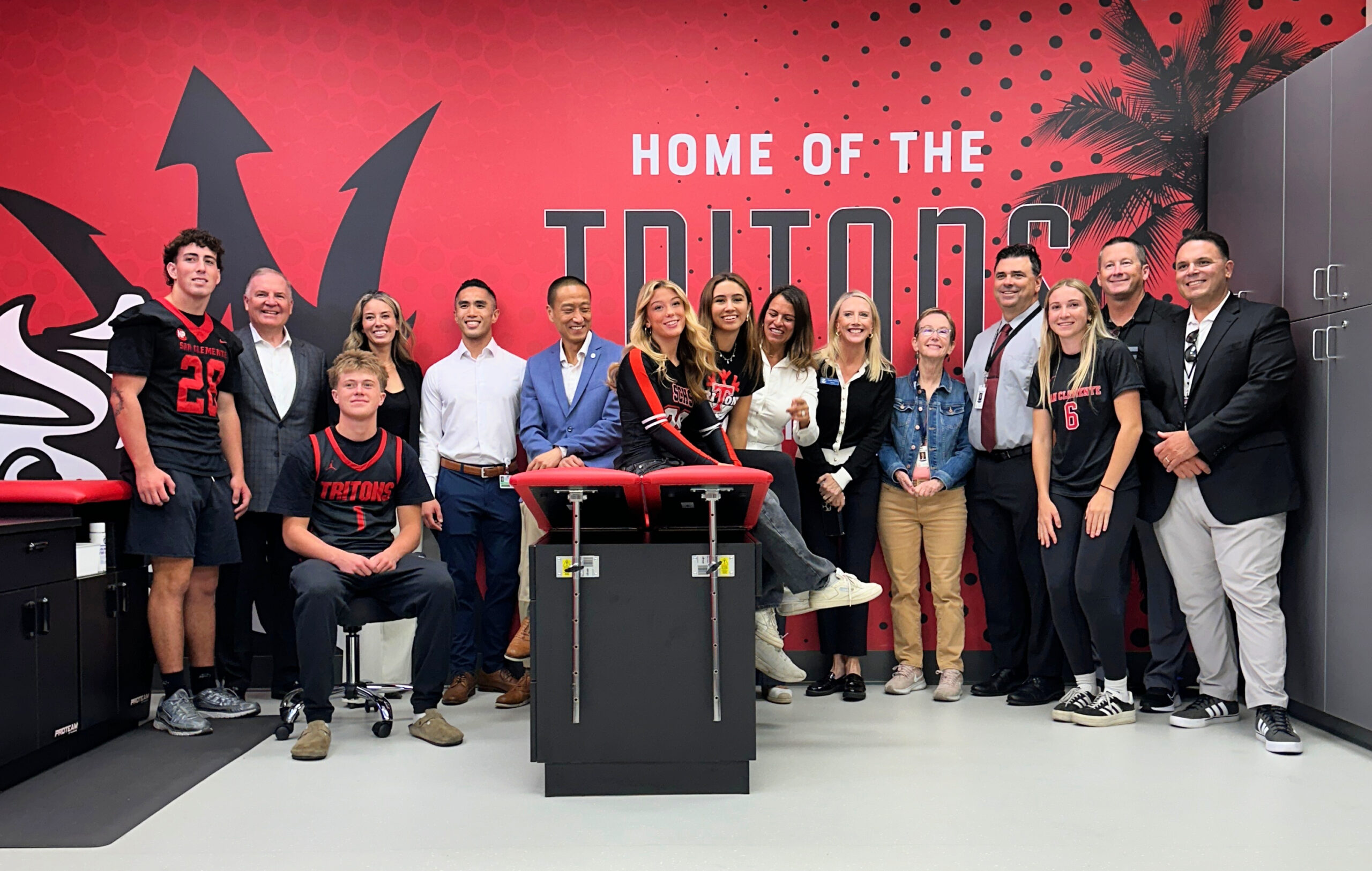 Group of San Clemente High School student-athletes and representatives from Hoag and Capistrano Unified School District standing and smiling inside the school’s new athletic training room, featuring a bright red wall with the words “Home of the Tritons.”