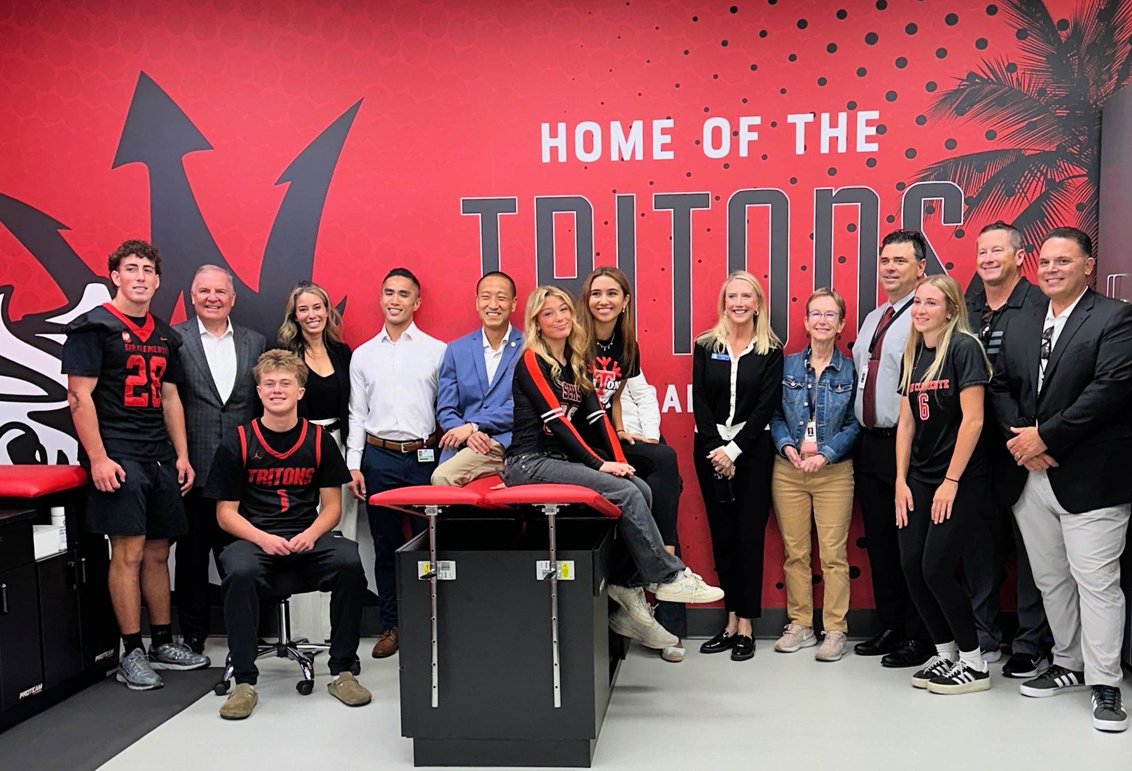 Group of San Clemente High School student-athletes and representatives from Hoag and Capistrano Unified School District standing and smiling inside the school’s new athletic training room, featuring a bright red wall with the words “Home of the Tritons.”