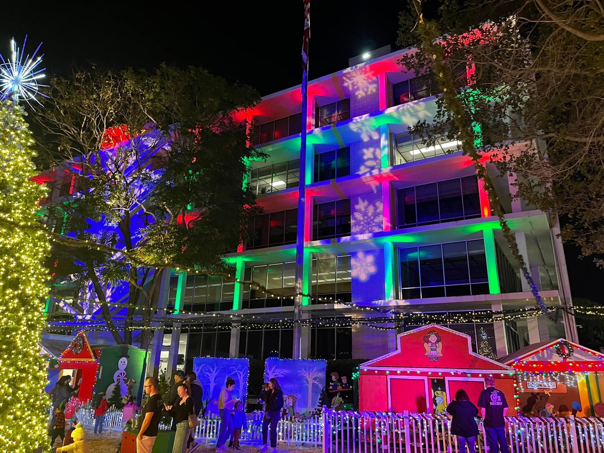 Families visit the Snoopy House display outside Costa Mesa City Hall during opening night in 2022, as colorful lights and snowflake projections illuminate the building. 