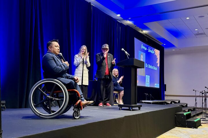 Dr. Stefan Bean is onstage holding a microphone as three event hosts stand and applaud behind him during the OC Influencers for Good Celebration; a screen beside the stage displays his name and award.