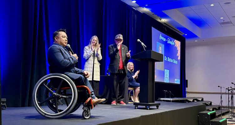 Dr. Stefan Bean is onstage holding a microphone as three event hosts stand and applaud behind him during the OC Influencers for Good Celebration; a screen beside the stage displays his name and award.