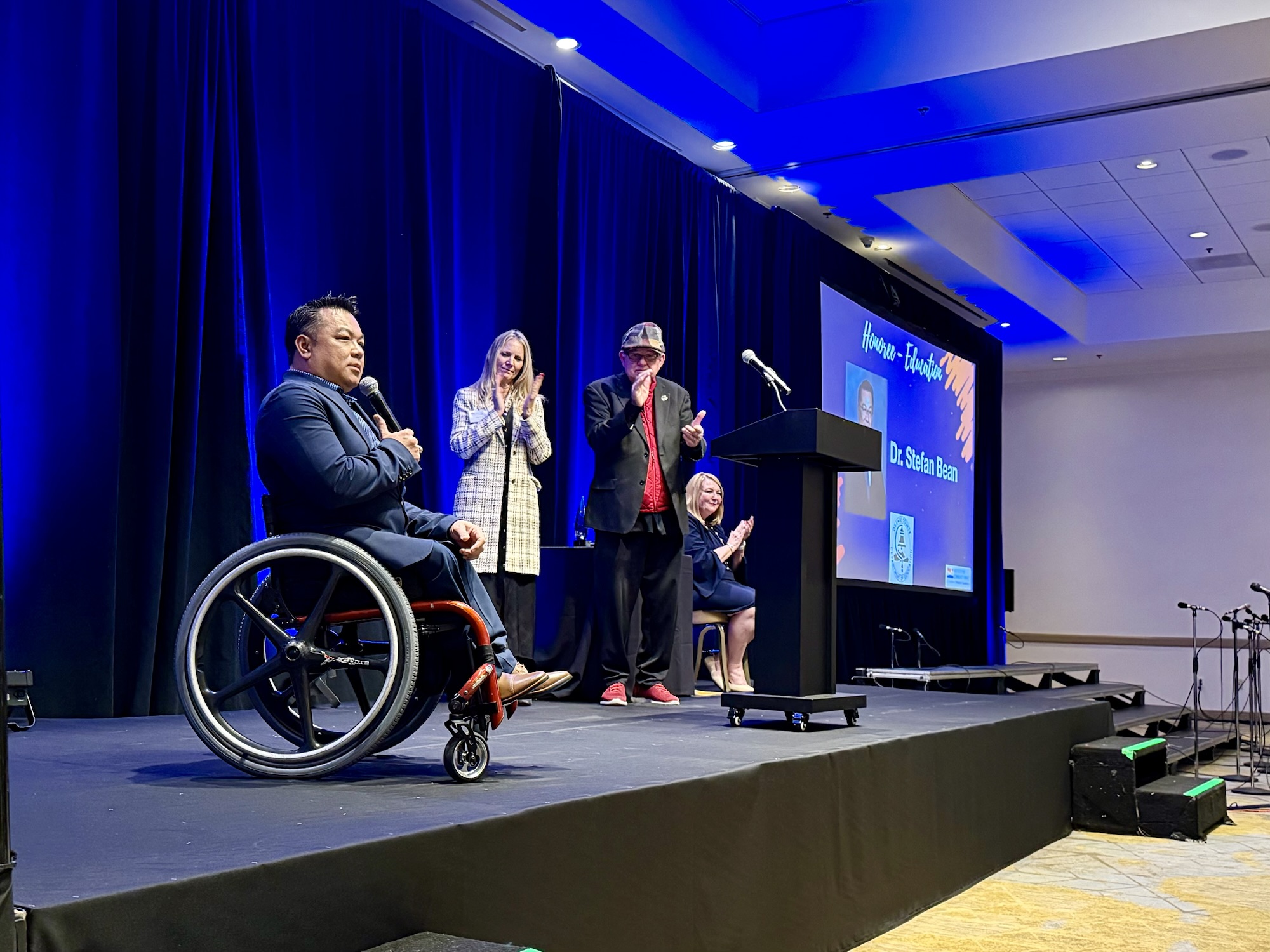 Dr. Stefan Bean is onstage holding a microphone as three event hosts stand and applaud behind him during the OC Influencers for Good Celebration; a screen beside the stage displays his name and award.