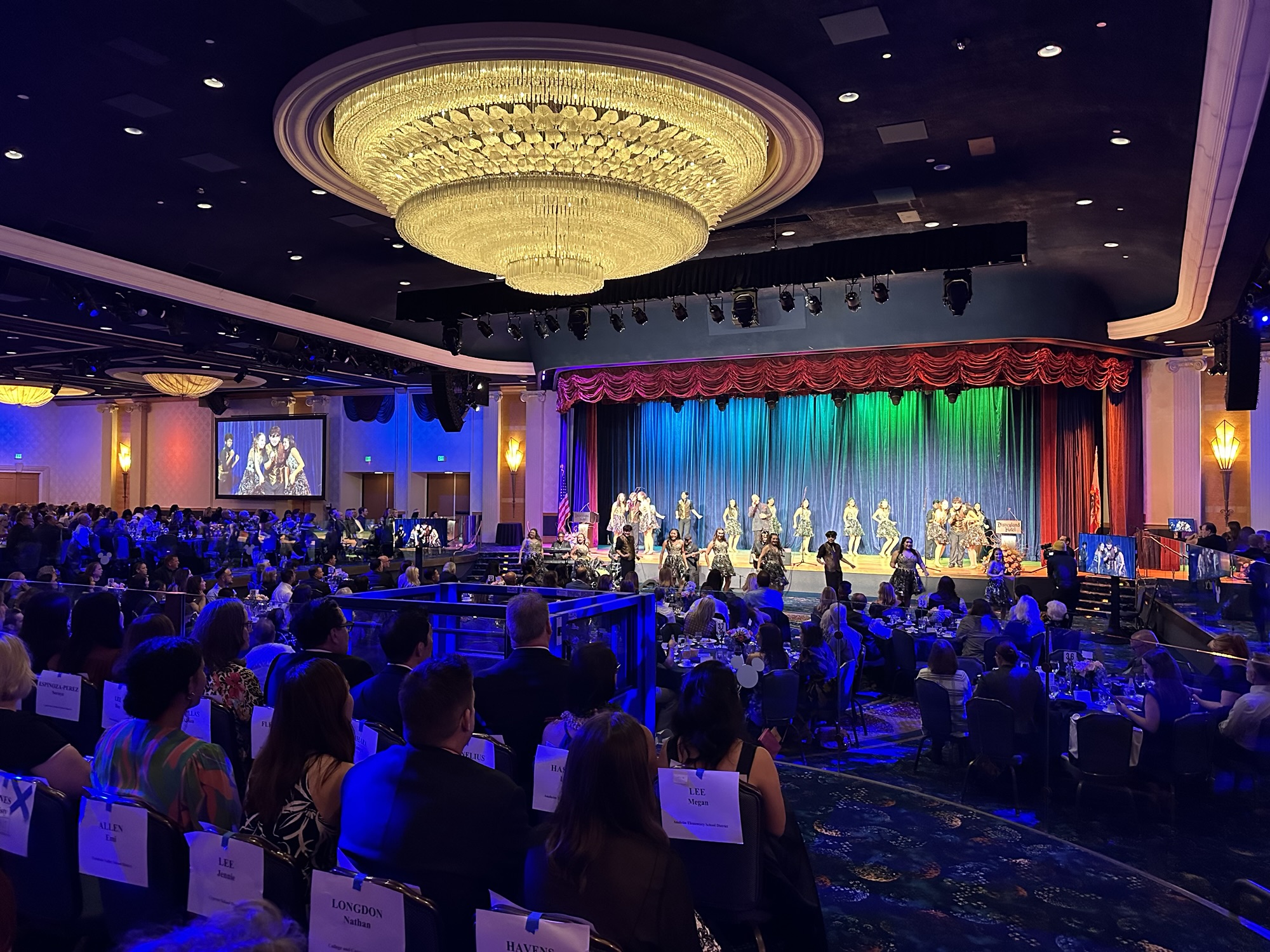 Brea Olinda High School’s Masquerade show choir performs on stage under colorful lighting during the 2026 Orange County Teachers of the Year gala at the Disneyland Hotel, with guests seated at round tables throughout the ballroom.