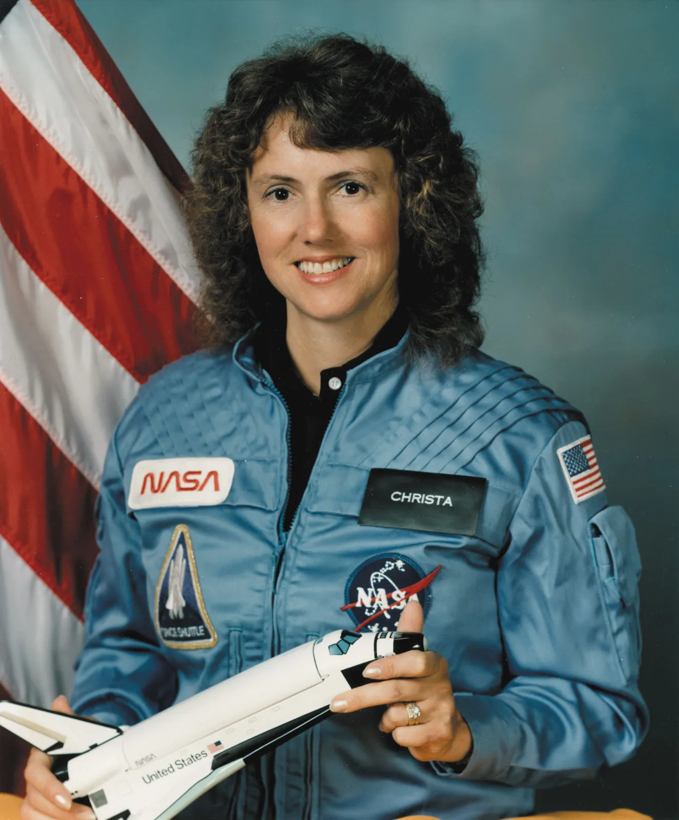 Christa McAuliffe wears a NASA flight suit and holds a model space shuttle in an official portrait taken before the Challenger mission.