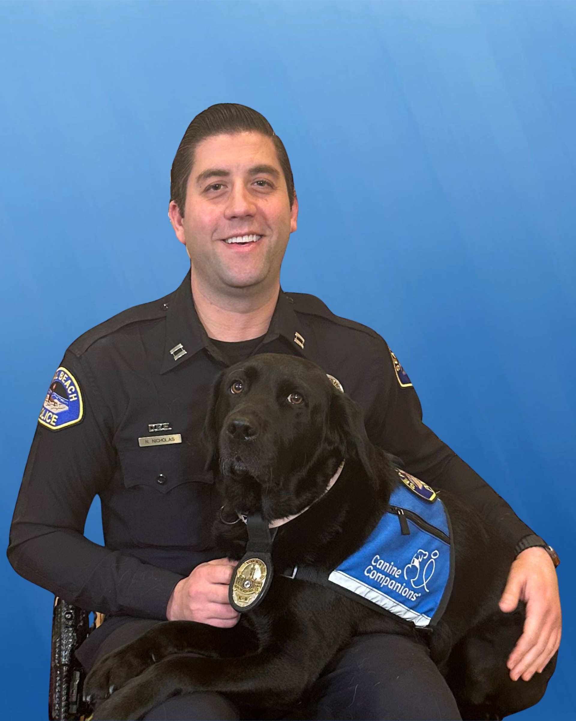 Seal Beach Police Capt. Nick Nicholas, in uniform, sits against a blue backdrop holding a black Labrador retriever wearing a blue “Canine Companions” service vest and medal.