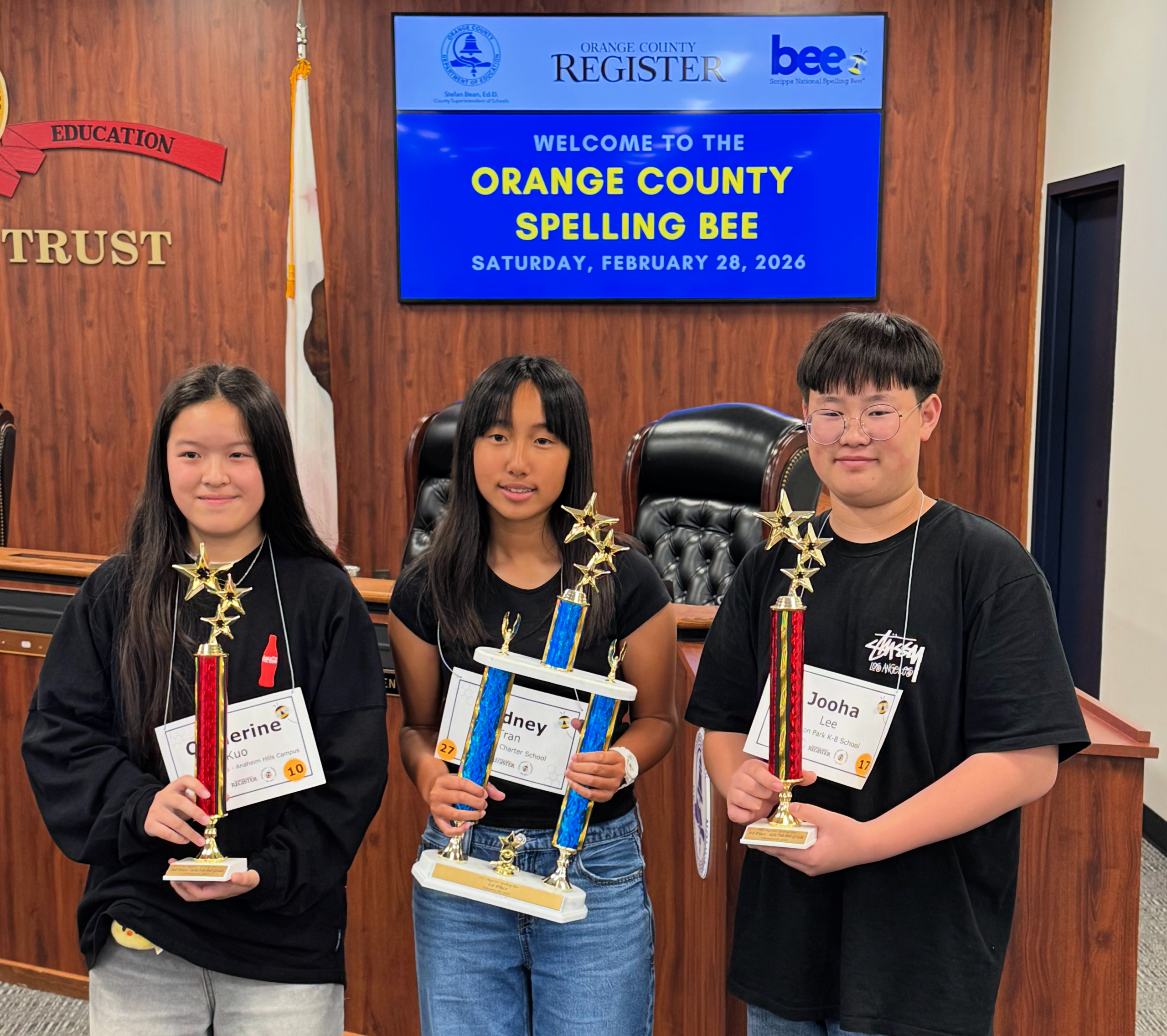 Sydney Tran, center, holds her first-place trophy alongside second-place finisher Catherine Kuo, left, and third-place finisher Jooha Lee following the 2026 Orange County Spelling Bee.
