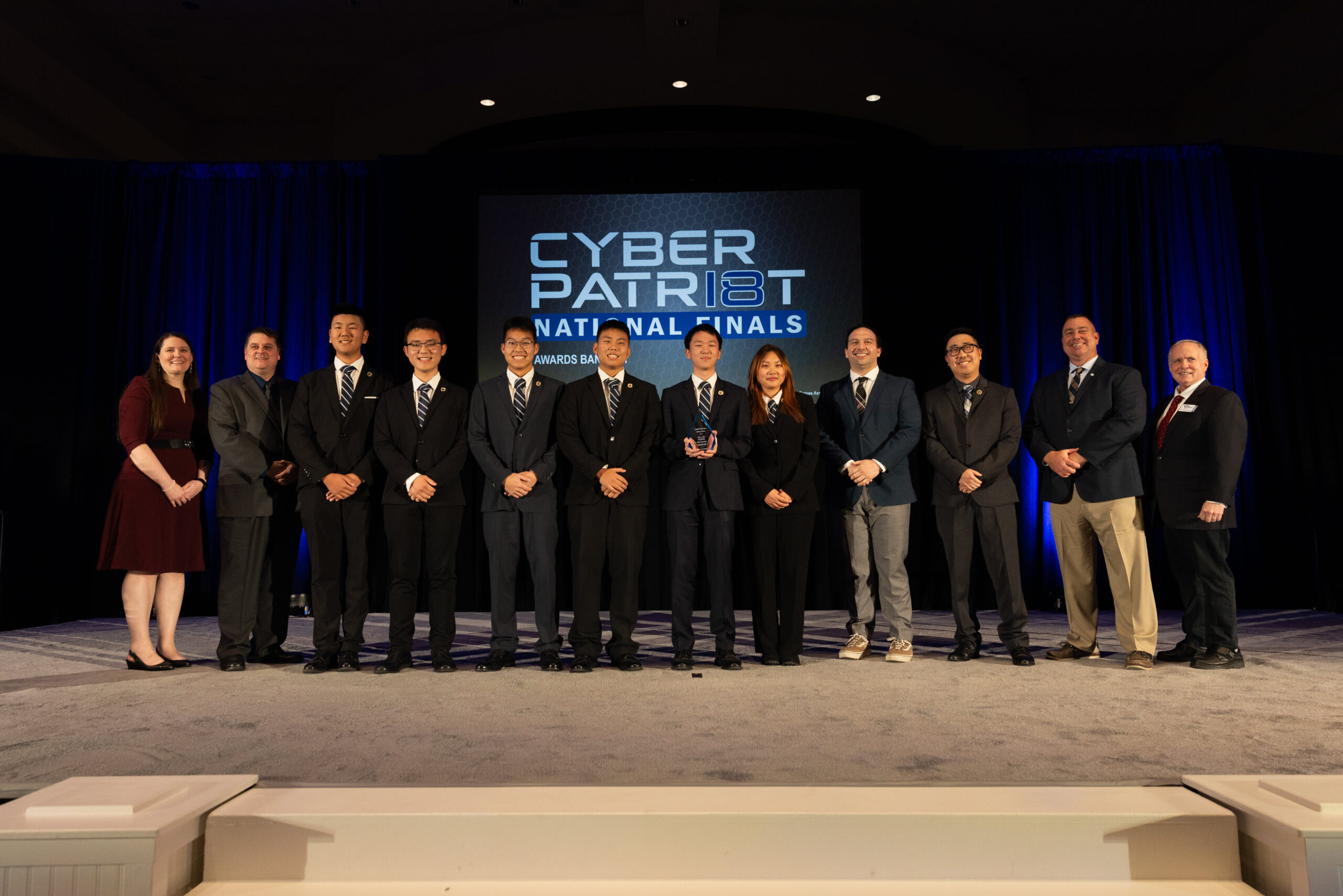 A group of Troy High School students and advisors stand on a stage at the CyberPatriot National Finals, posing with an award in front of a large screen displaying “CyberPatriot National Finals.”