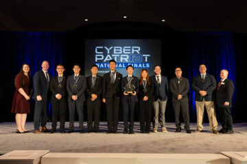 A group of students and advisors from Troy High School stand on a stage at the CyberPatriot National Finals, holding an award in front of a large screen displaying “CyberPatriot National Finals.”