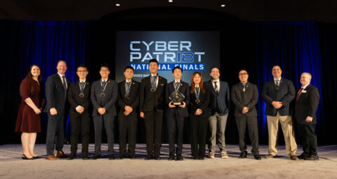 A group of students and advisors from Troy High School stand on a stage at the CyberPatriot National Finals, holding an award in front of a large screen displaying “CyberPatriot National Finals.”