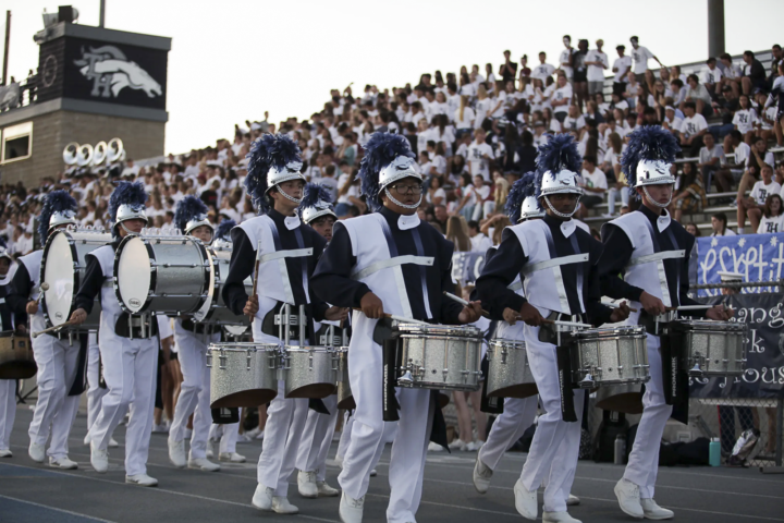 Students in Trabuco Hills High School’s marching band drumline, wearing navy and white uniforms with plumed helmets, perform during a football game in front of a packed stadium crowd.