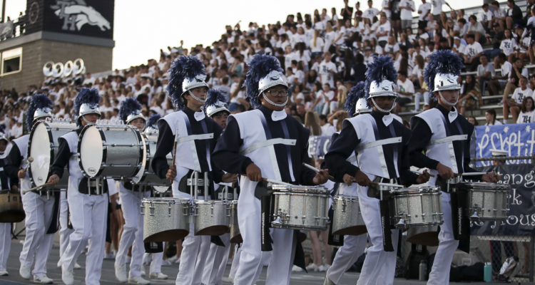 Students in Trabuco Hills High School’s marching band drumline, wearing navy and white uniforms with plumed helmets, perform during a football game in front of a packed stadium crowd.